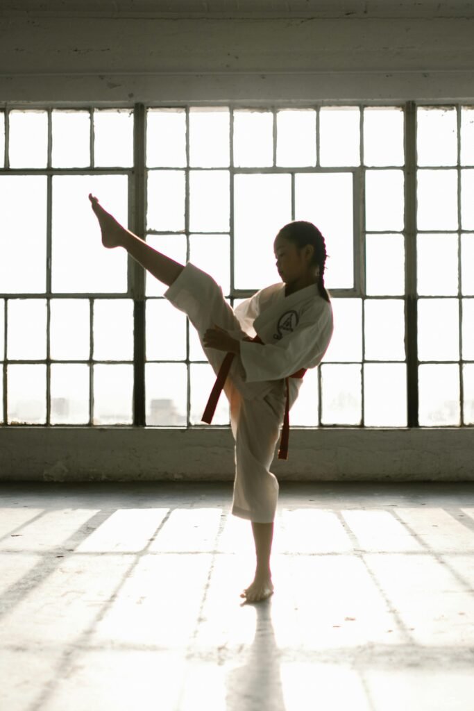 A young girl in a martial arts uniform performing a high kick in a sunlit studio.