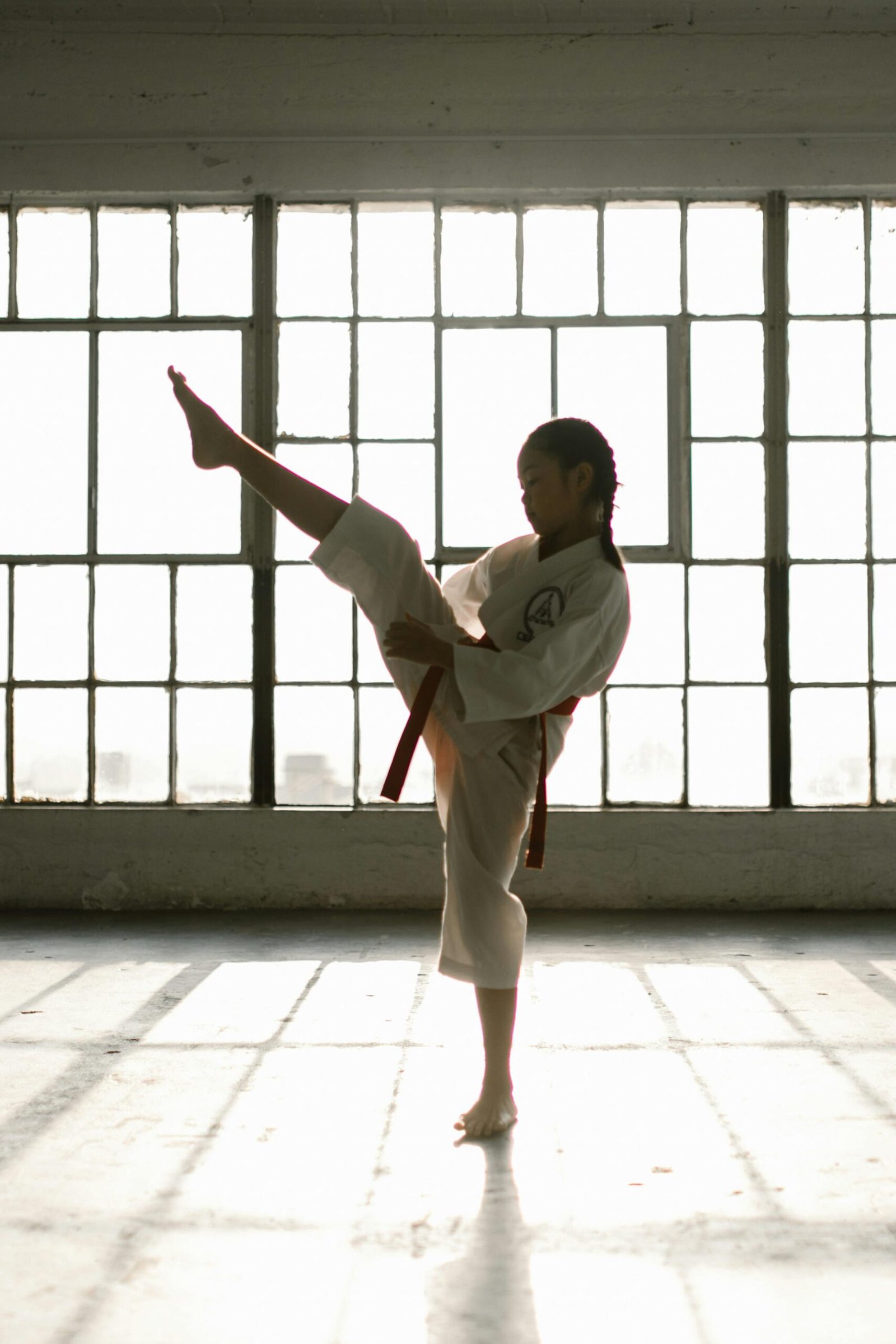 A young girl in a martial arts uniform performing a high kick in a sunlit studio.