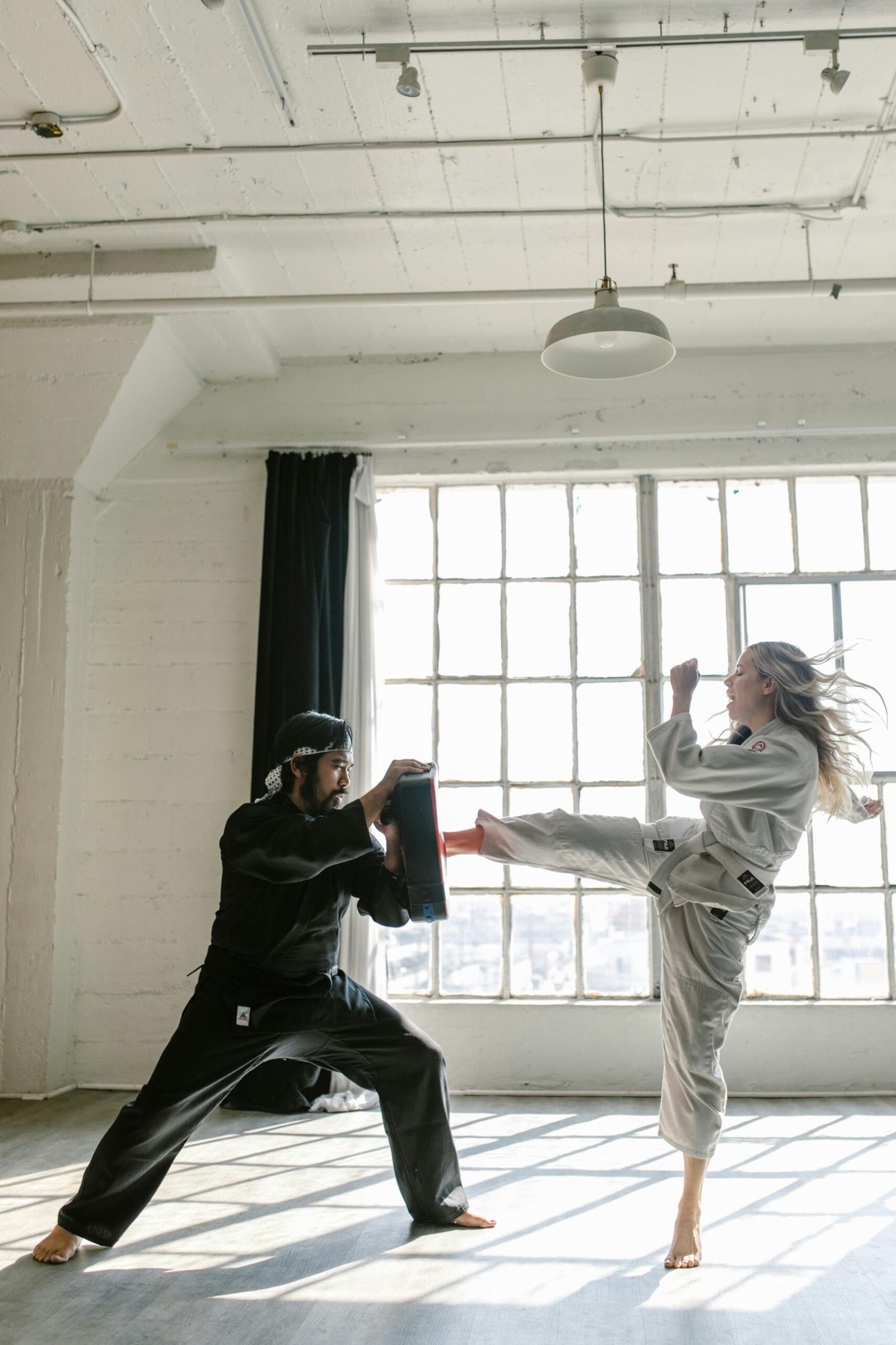 Martial artists practicing kicks and defense techniques indoors in a sunlight gym.